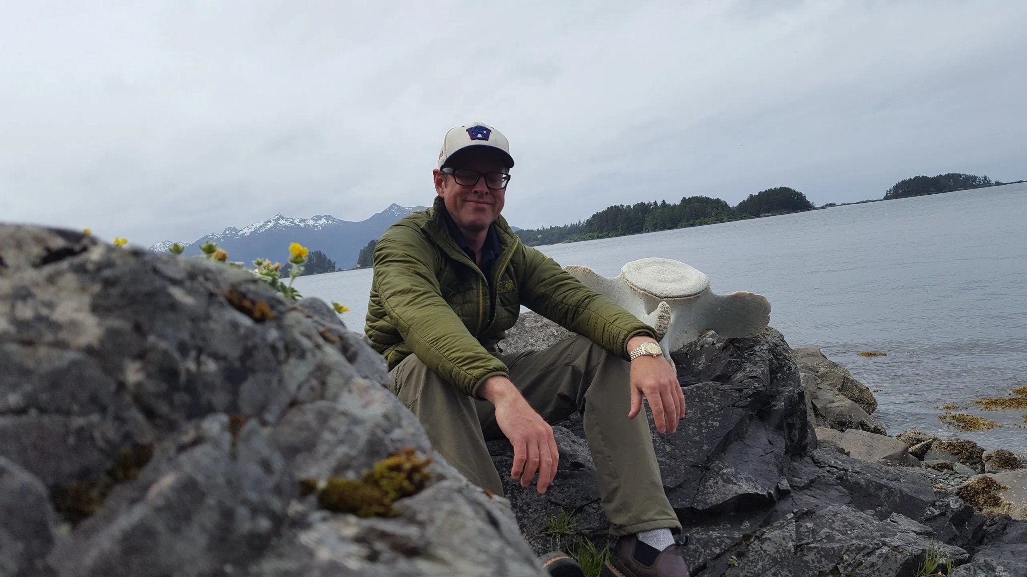 Capt. Xander sitting on coastal rocks with a whale vertebra and Sitka Sound behind him